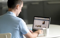 Homem de costas sentado à mesa, trabalhando em um notebook com uma xícara de café ao lado.