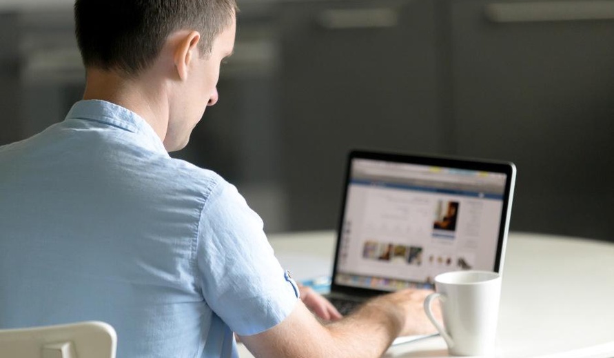 Homem de costas sentado à mesa, trabalhando em um notebook com uma xícara de café ao lado.